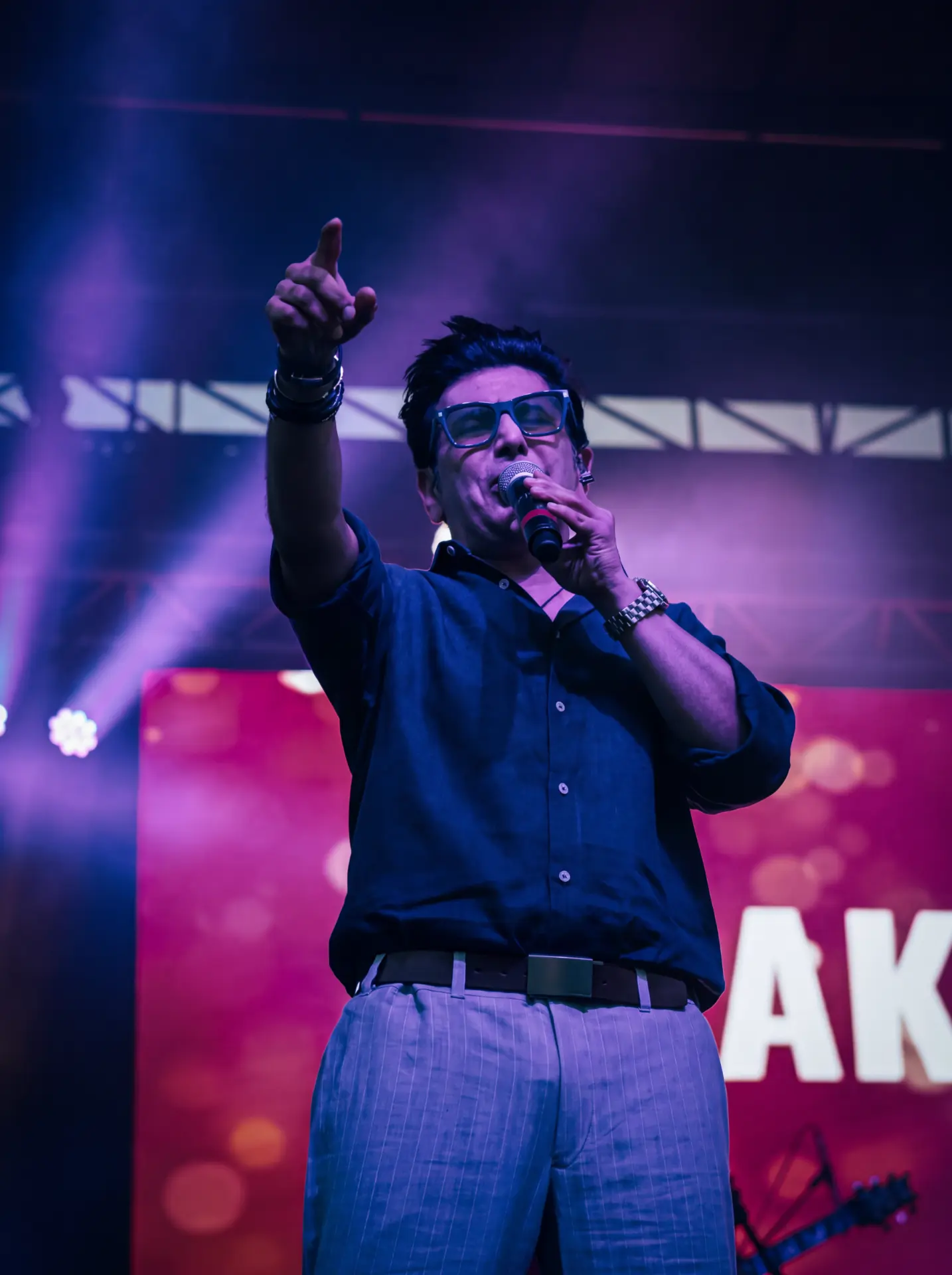 Singer in blue glasses pointing to the crowd under purple stage lights