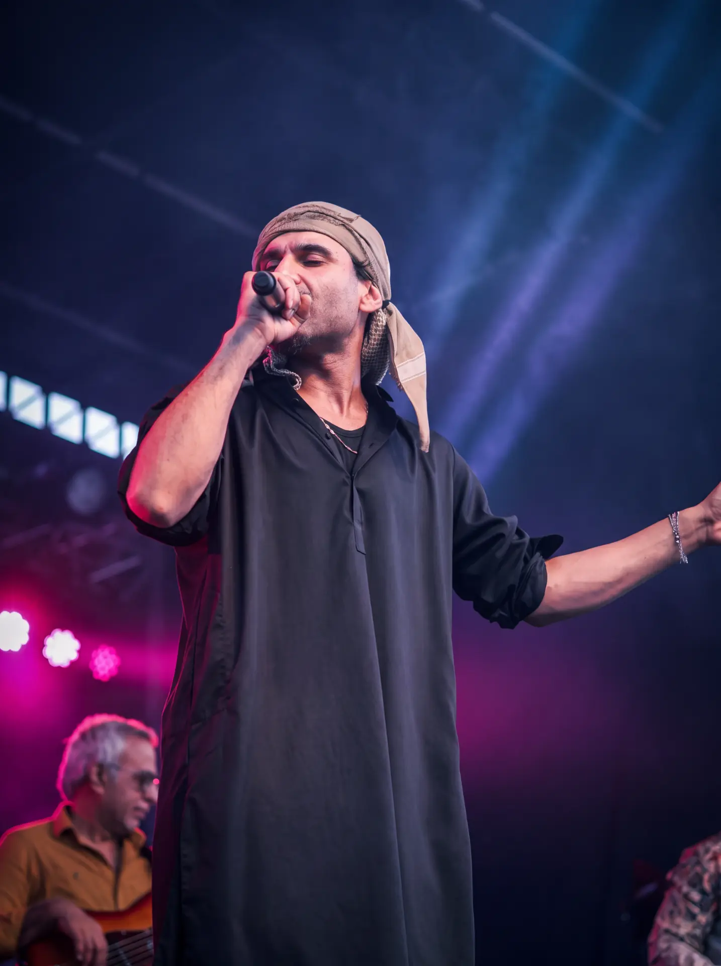 Singer in a black robe and headwrap performing beneath blue and pink stage lights