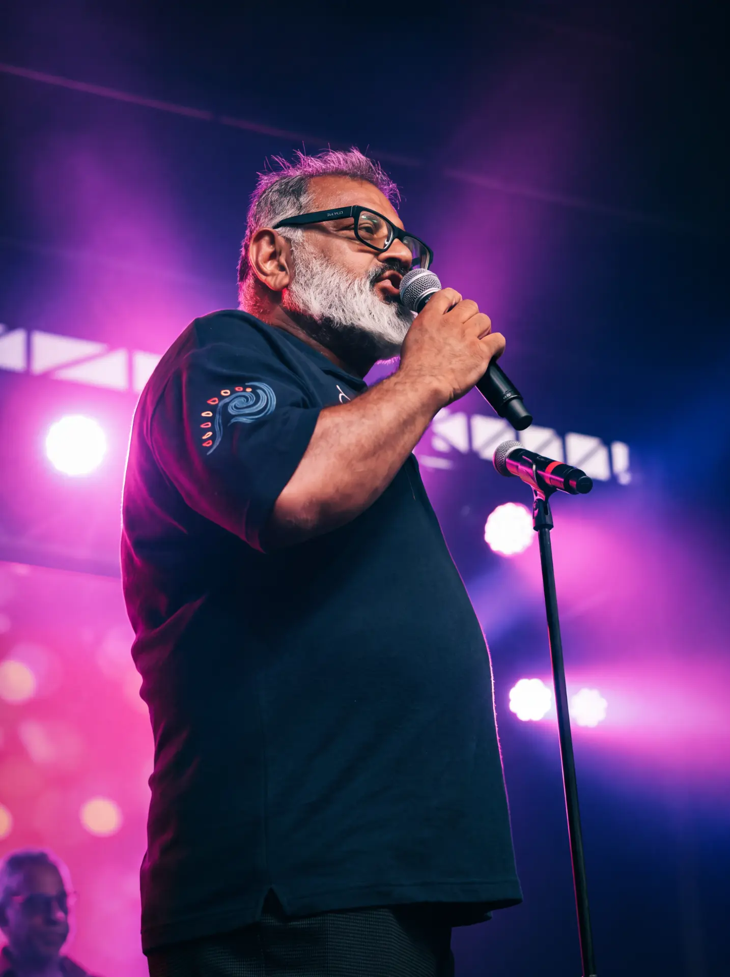 Vocalist with a grey beard and glasses singing under pink stage lights