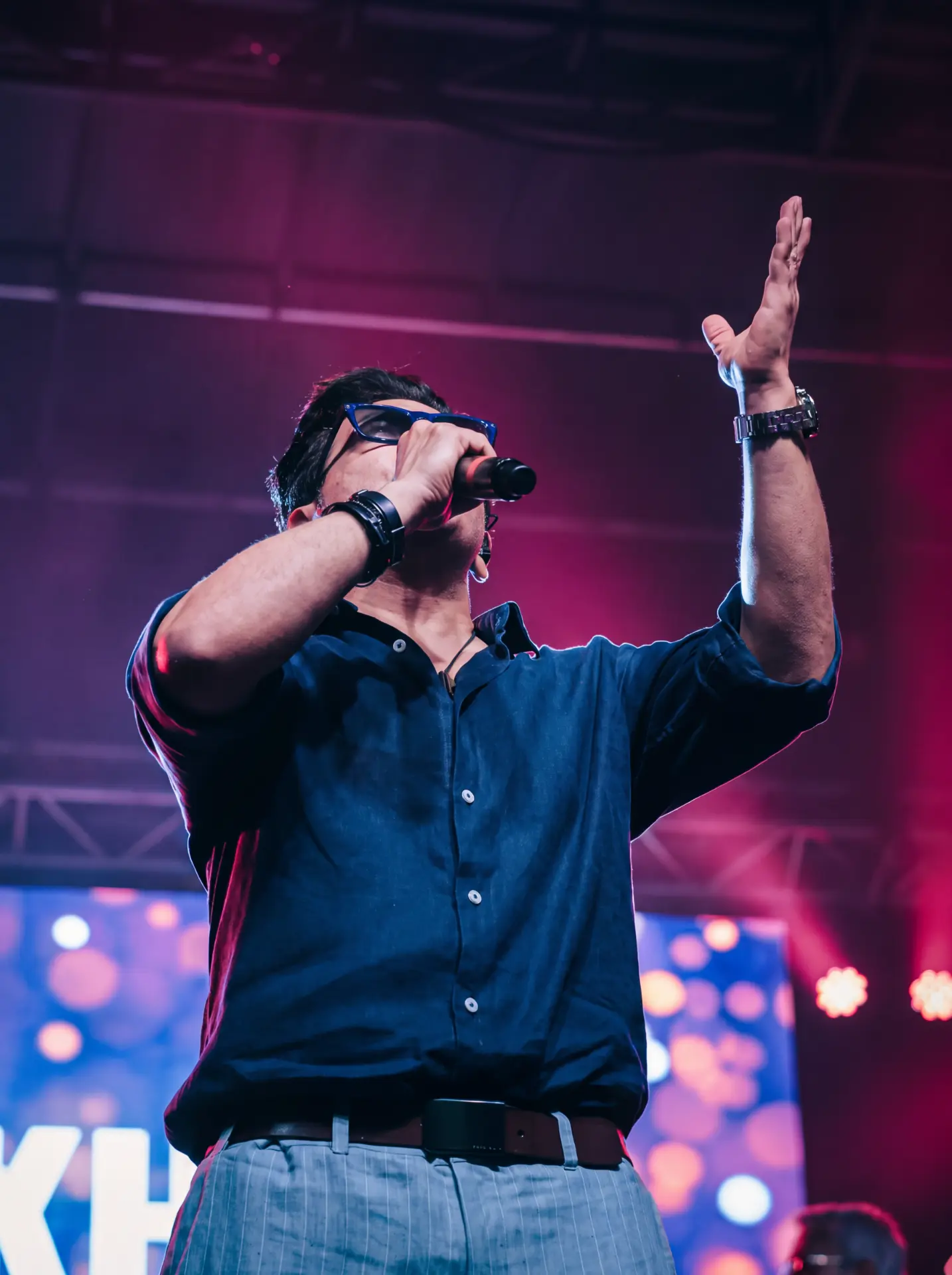 Singer in blue glasses with a hand raised under magenta lights